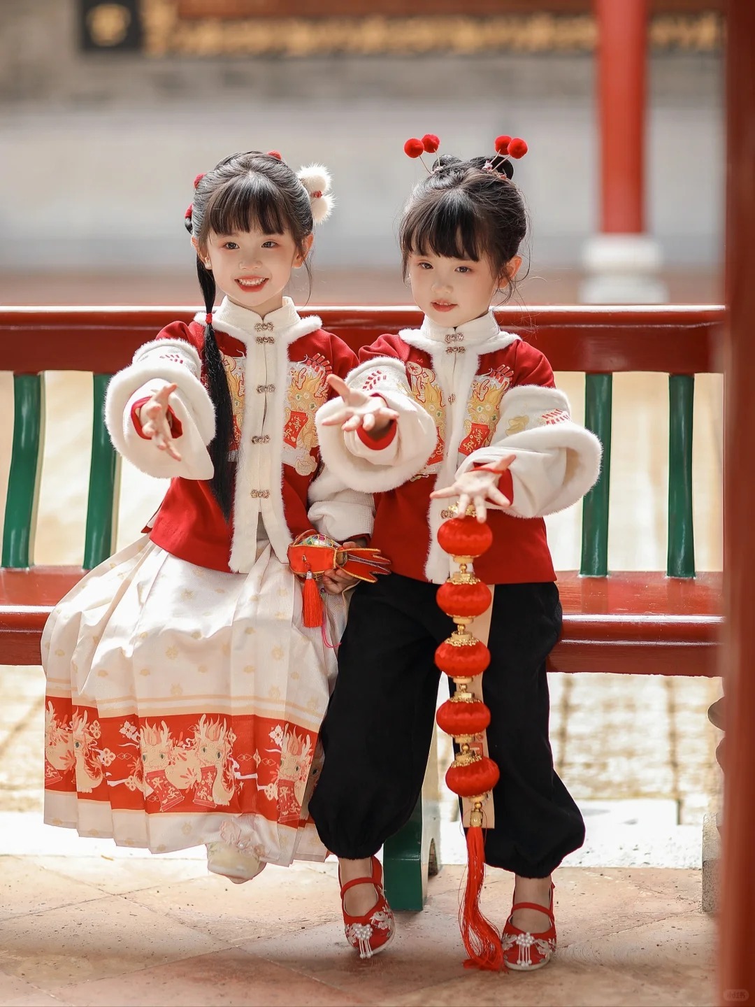 Two young girls wearing festive Chinese New Year clothes with traditional red and white designs, holding decorative lantern tassels.