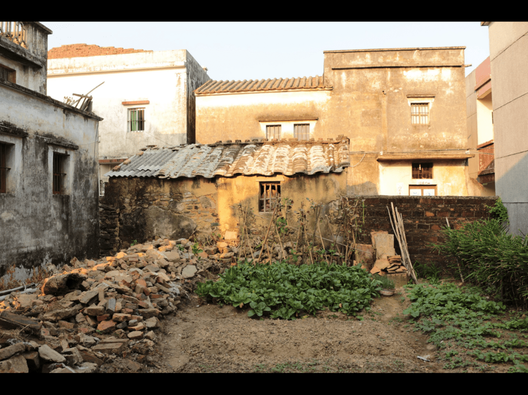 An empty lot in the Chan ancestral village of Lianjiangli, where Grandpa Chan’s house once stood, now surrounded by rubble, plants, and neighboring structures.