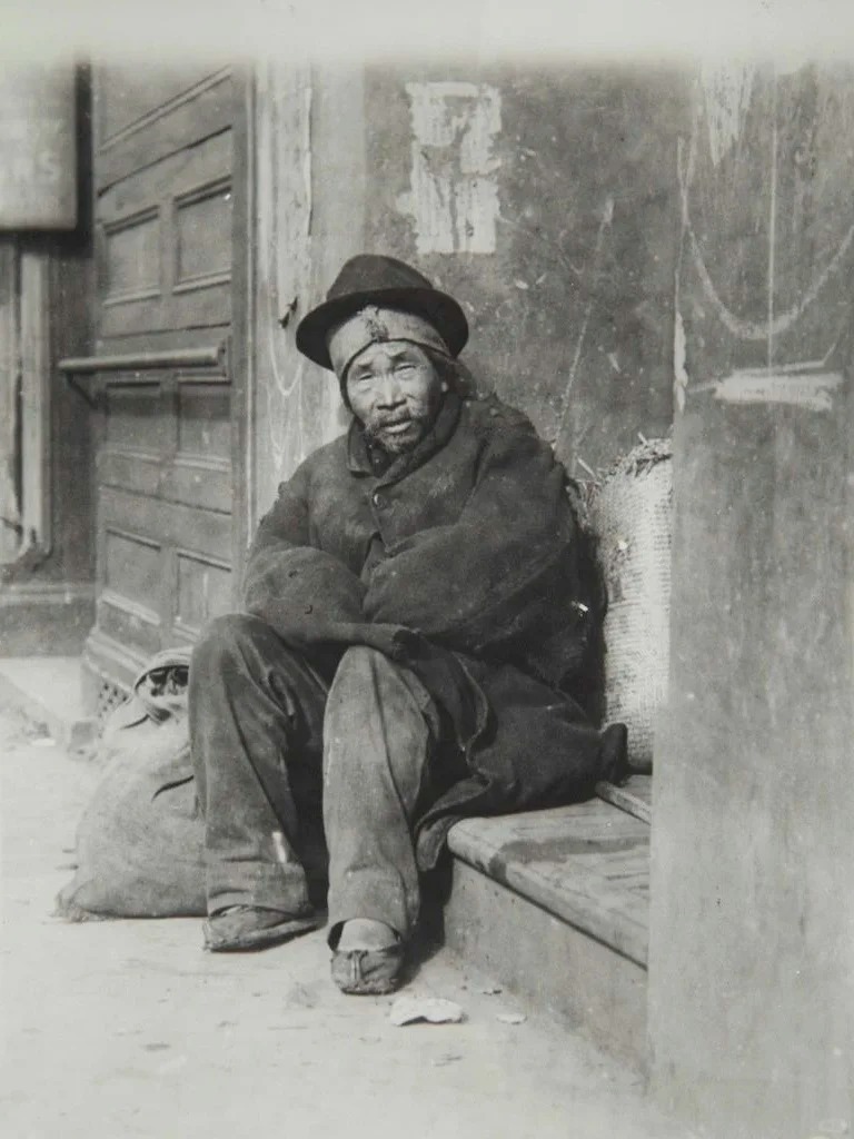 A seated Wuyi migrant man with worn clothing and belongings beside him, representing the economic hardships and social isolation faced by early Chinese immigrants.