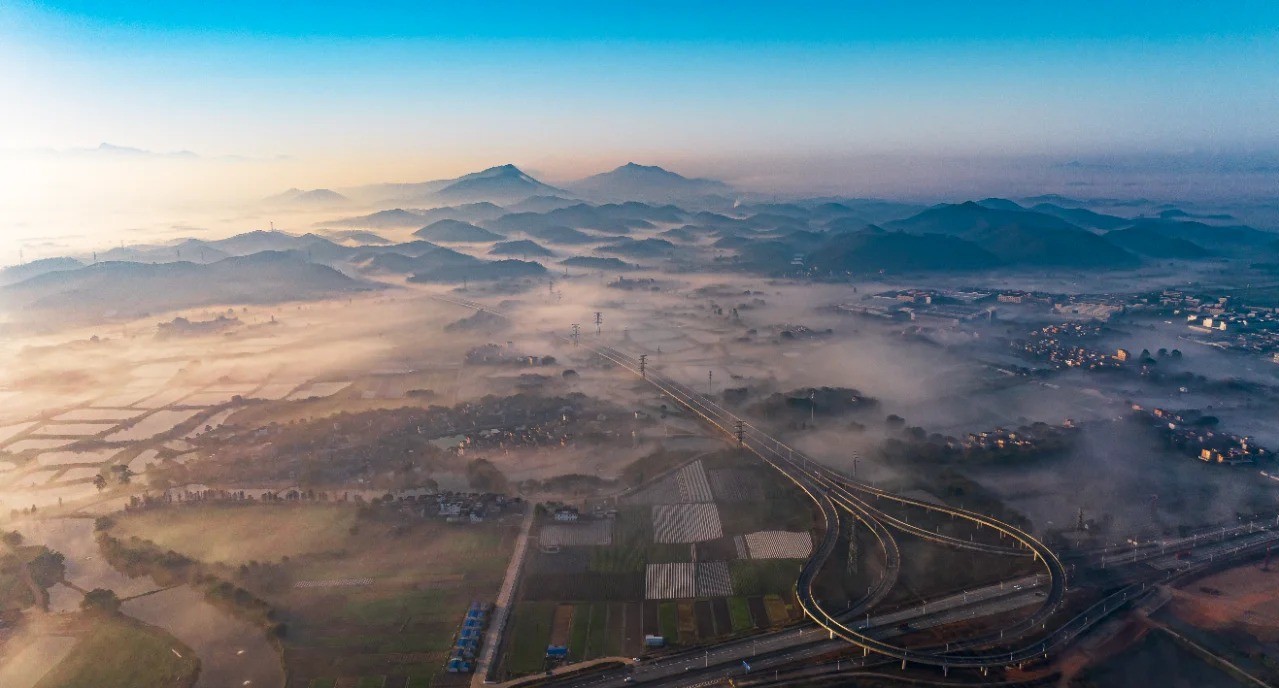 An aerial view of Kaiping in Guangdong Province, showing a misty landscape with rolling hills, agricultural fields, villages, and modern infrastructure like highways and railways under a clear blue sky.