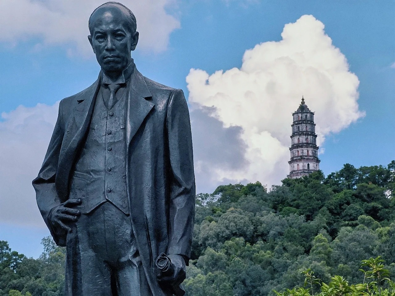 A statue of Liang Qichao in Xinhui stands in front of a lush green landscape with a traditional pagoda and a partly cloudy sky.