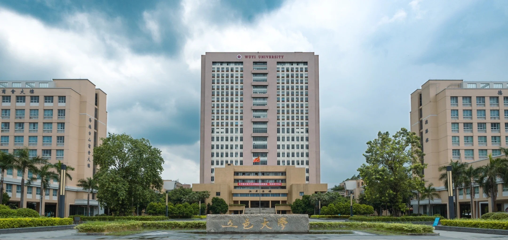 A photograph of Wuyi University in Guangdong Province, featuring a tall central building with the university’s name at the top, flanked by two smaller buildings, surrounded by palm trees, greenery, and a stone sign with Chinese characters under a cloudy sky.