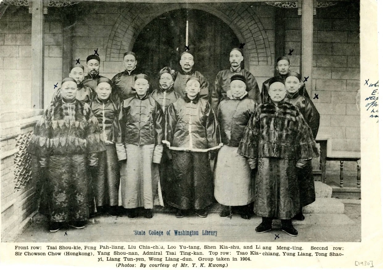 A black-and-white photograph of a group of men dressed in traditional Chinese attire, standing in front of an arched doorway. The individuals appear to be part of a formal gathering, with some wearing elaborate robes and others in more modest clothing.