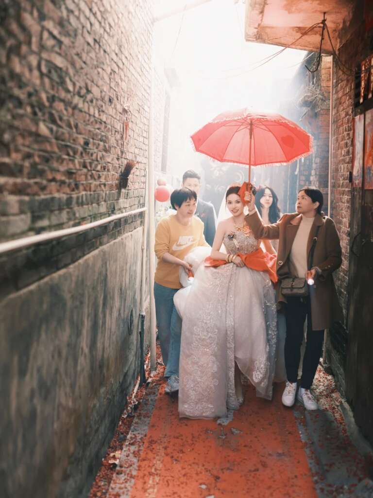 Bride walking through village alley with red umbrella, merging tradition and modern intention