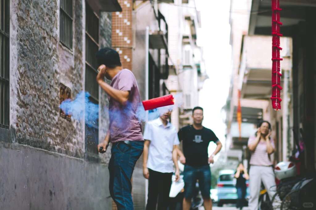 A man lights firecrackers in a narrow alleyway of a rural village in China, with smoke and sparks filling the air as neighbors watch.